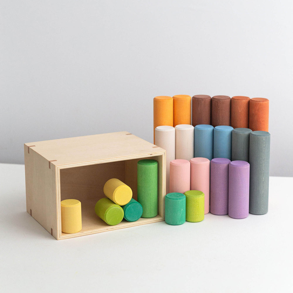 A set of colorful wooden cylinder blocks in various sizes, displayed both inside and outside of a wooden box. The cylinders are in multiple shades including green, yellow, blue, pink, orange, and gray.