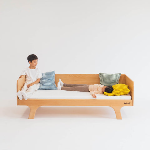 A child sitting on a beige Hug Hug Bed 90x200 with a Teenage Boy lying down on it, surrounded by pillows of different colors.