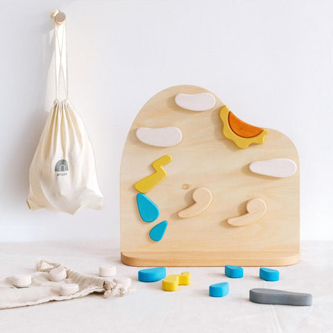 A wooden weather board toy with metal board and magnetic weather pieces, including clouds, sun, and rain symbols, displayed on a white background with a few other toys and a fabric bag in the background.