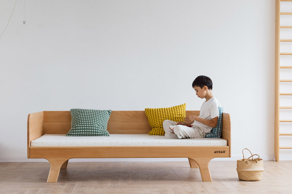 A child leaning on a beige Hug Hug Bed 90x200 , surrounded by pillows of different colors.