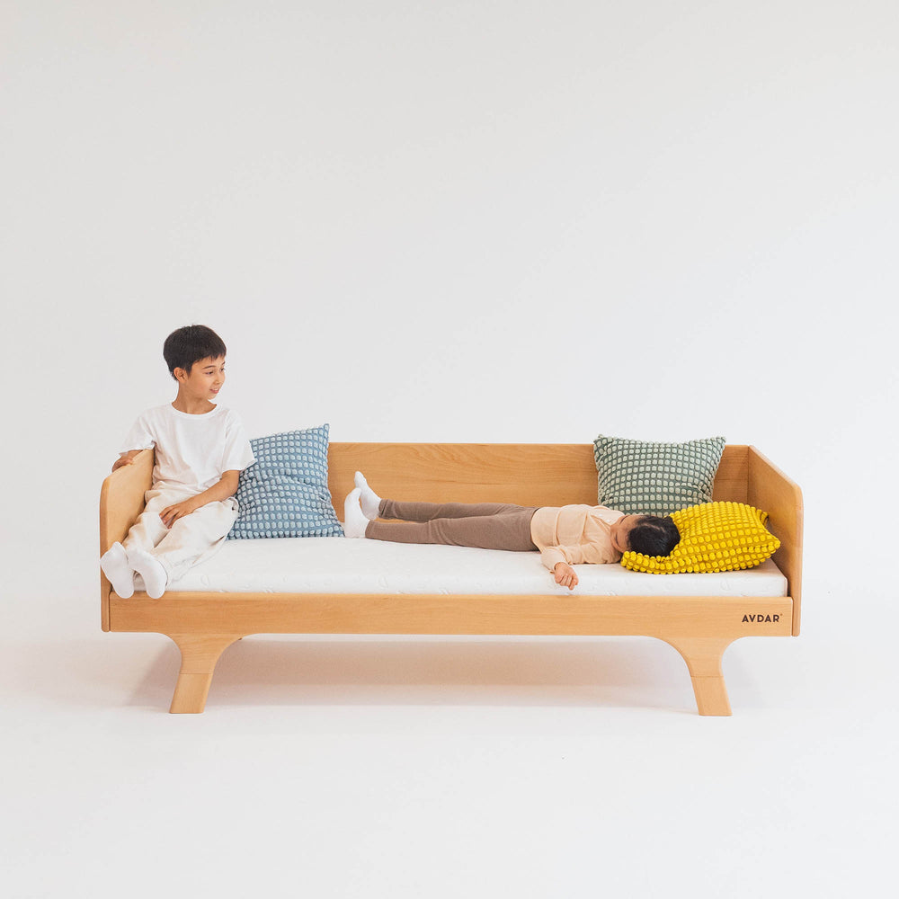 A child sitting on a beige Hug Hug Bed 90x200 with a Teenage Boy lying down on it, surrounded by pillows of different colors.